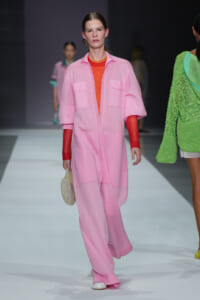 Model walks the runway in a loose pink jumpsuit layered over an orange top, with red sleeves and a small beaded bag.