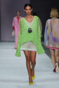 Model walks the runway in a lime-green knitted mesh sweater over a sheer white dress, accessorized with a black pendant necklace and yellow flats.