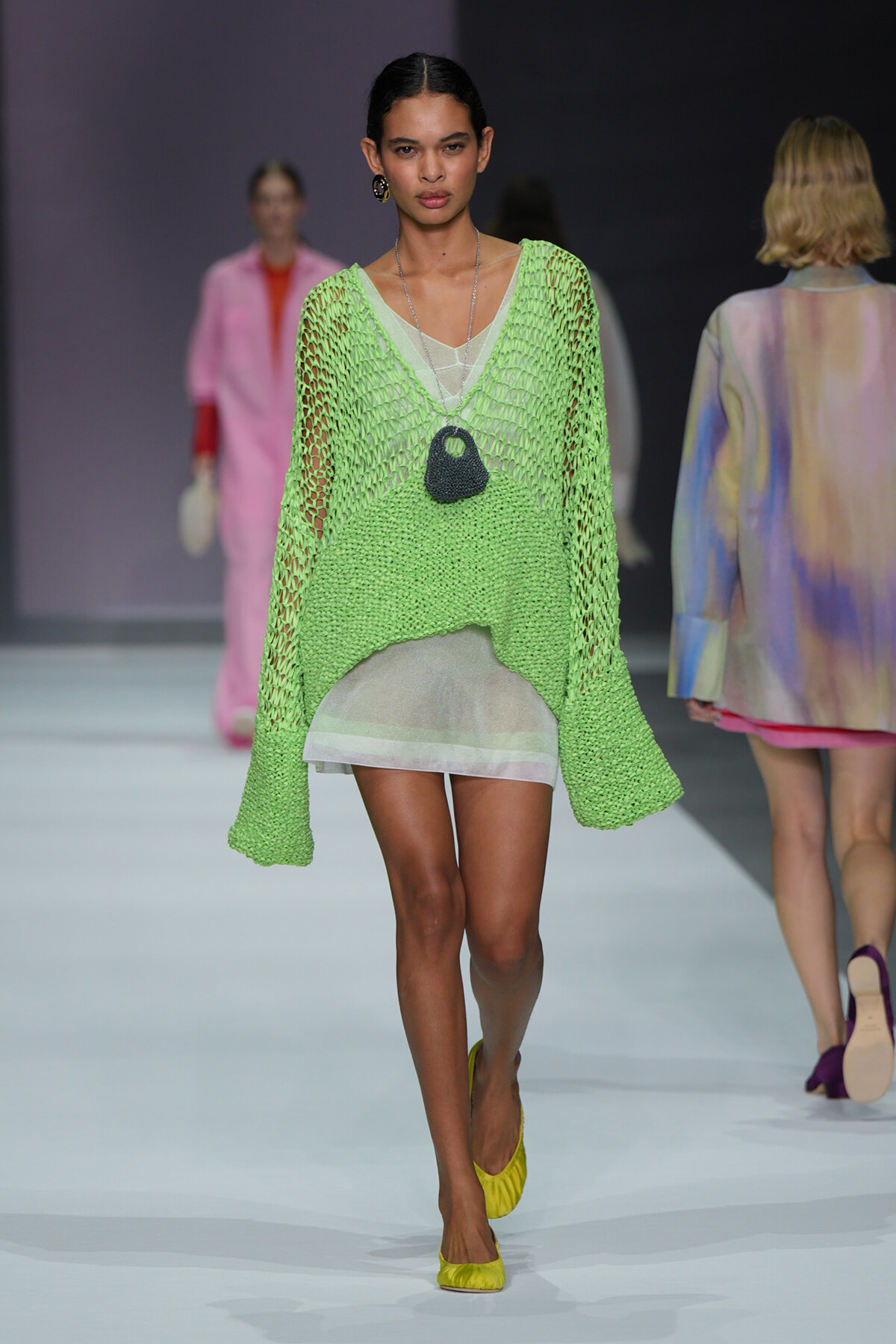 Model walks the runway in a lime-green knitted mesh sweater over a sheer white dress, accessorized with a black pendant necklace and yellow flats.