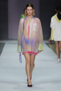 Model walking the runway in a pastel gradient oversized shirt with pink shorts, holding a lavender bag and wearing purple flats.
