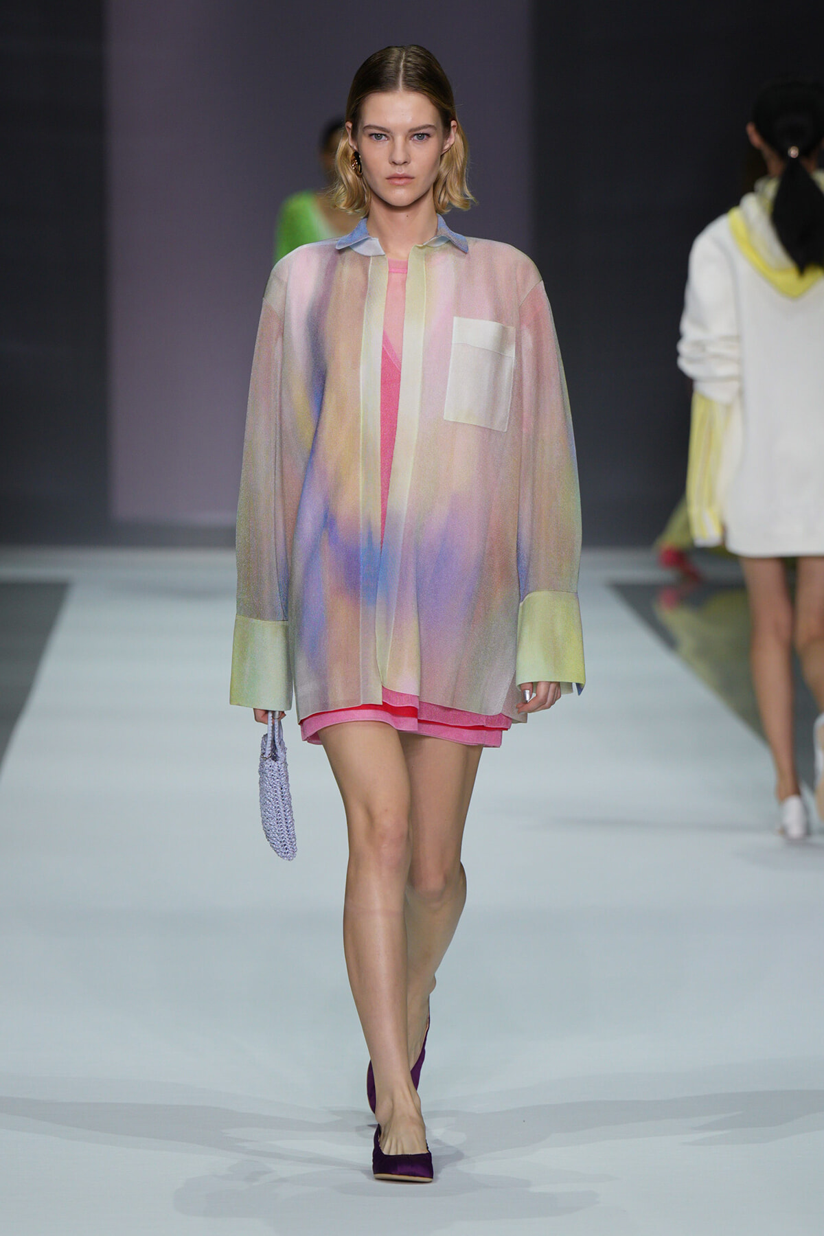 Model walking the runway in a pastel gradient oversized shirt with pink shorts, holding a lavender bag and wearing purple flats.