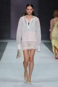 Model walking the runway in a sheer white tunic with mint-green trim and floral embroidery, carrying a beaded clutch.