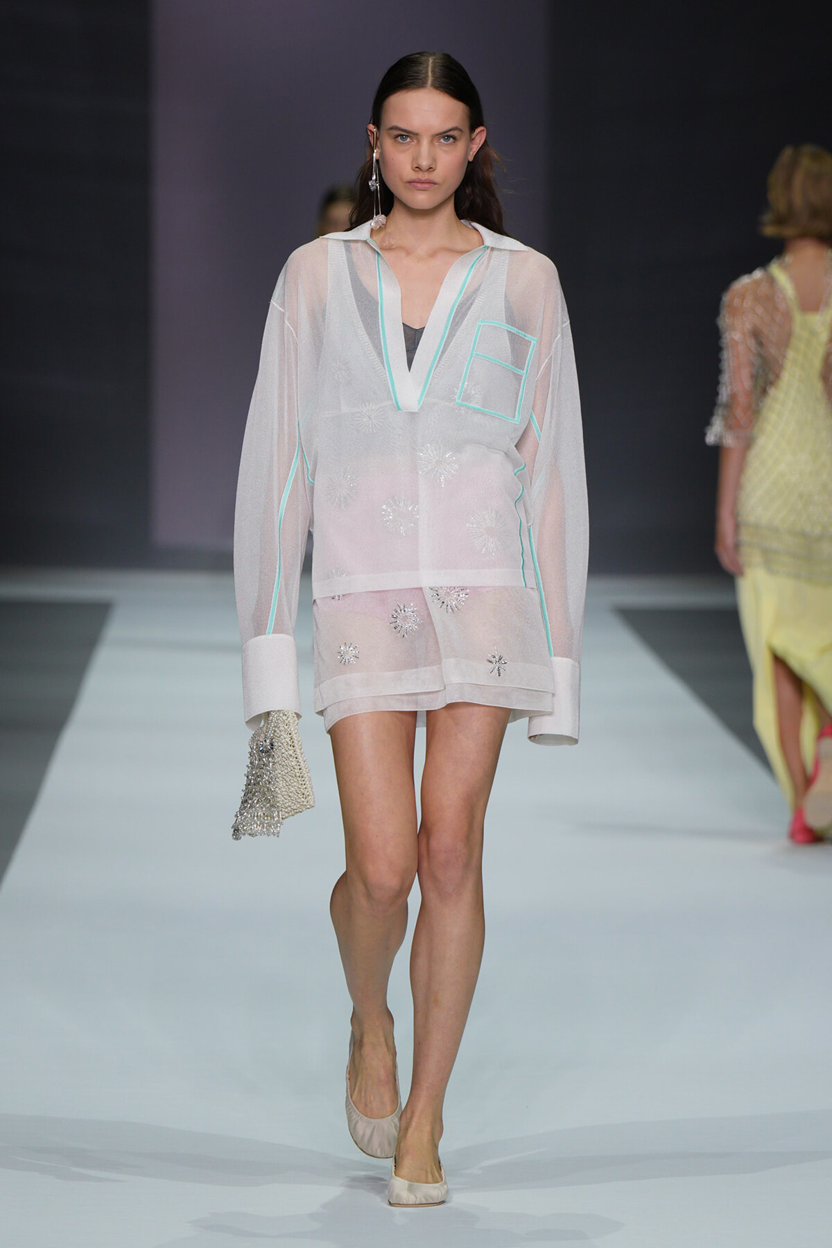 Model walking the runway in a sheer white tunic with mint-green trim and floral embroidery, carrying a beaded clutch.