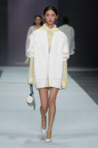Runway model in an oversized white zip-front coat with pale yellow striped sleeves and a yellow hoodie underneath, carrying a small round bag.