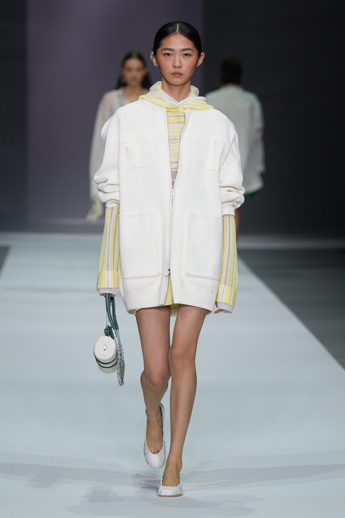 Runway model in an oversized white zip-front coat with pale yellow striped sleeves and a yellow hoodie underneath, carrying a small round bag.