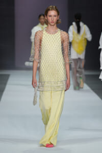 Runway model in a pale yellow tunic with a silver crystal-net overlay and matching wide-leg pants; pink shoes.