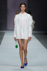 Model on a runway in a white shirt dress with mint piping, bright blue heels, and a small green beaded handbag.