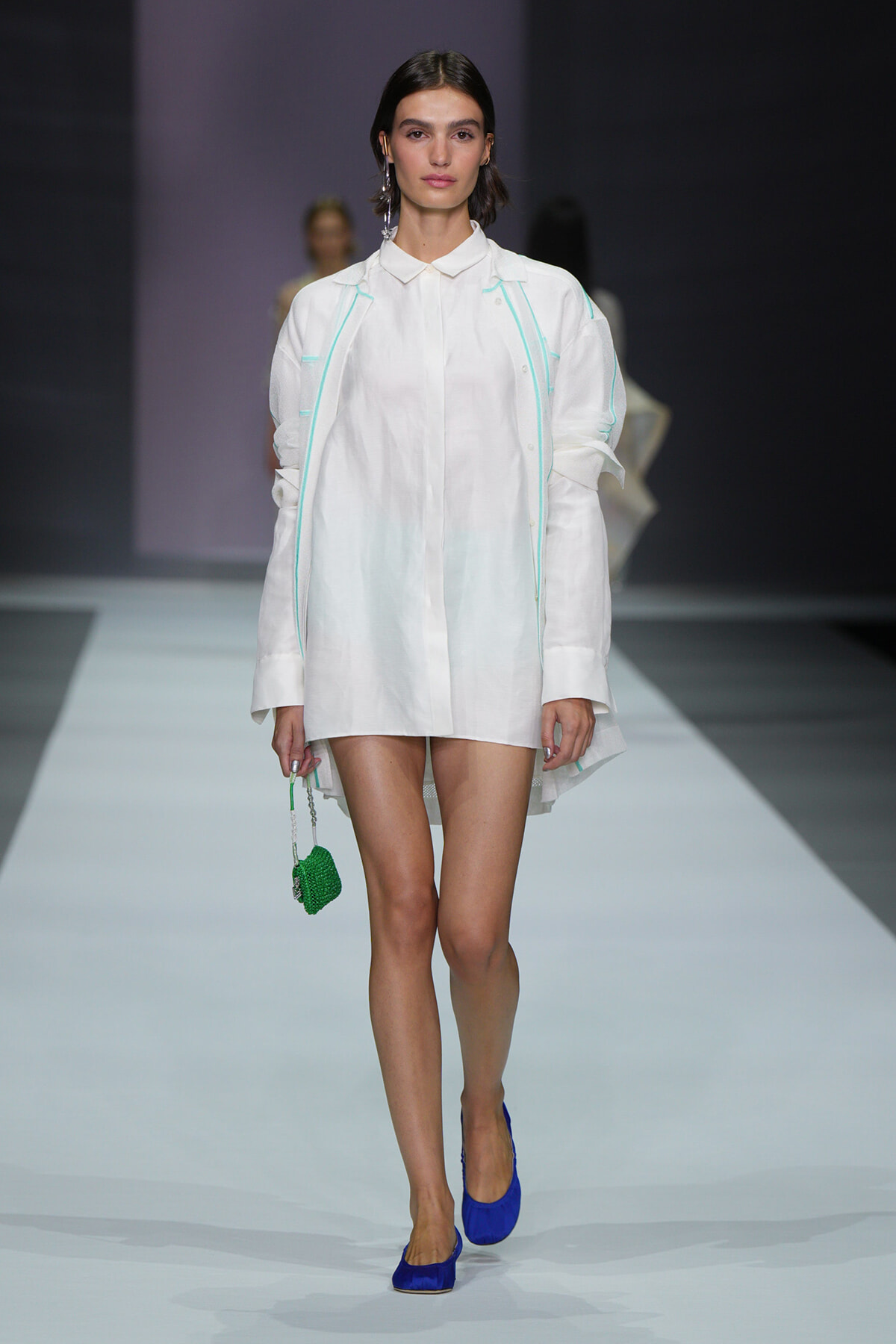 Model on a runway in a white shirt dress with mint piping, bright blue heels, and a small green beaded handbag.