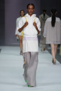 Model on a fashion runway wearing a white zip-up hoodie with a lavender waistband and oversized gray cargo pants, carrying a yellow net bag.