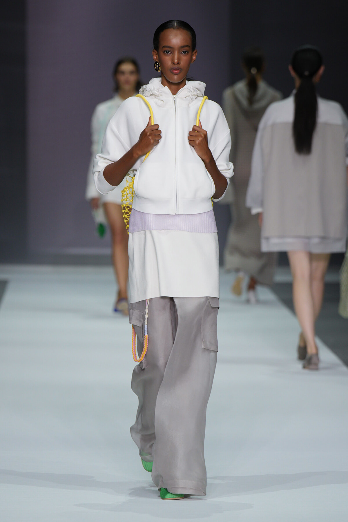 Model on a fashion runway wearing a white zip-up hoodie with a lavender waistband and oversized gray cargo pants, carrying a yellow net bag.
