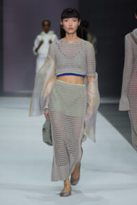 Model walks a fashion runway wearing a gray perforated knit crop top and matching skirt with neon lime and blue underlayers and sheer cape sleeves, carrying a glittery silver handbag.