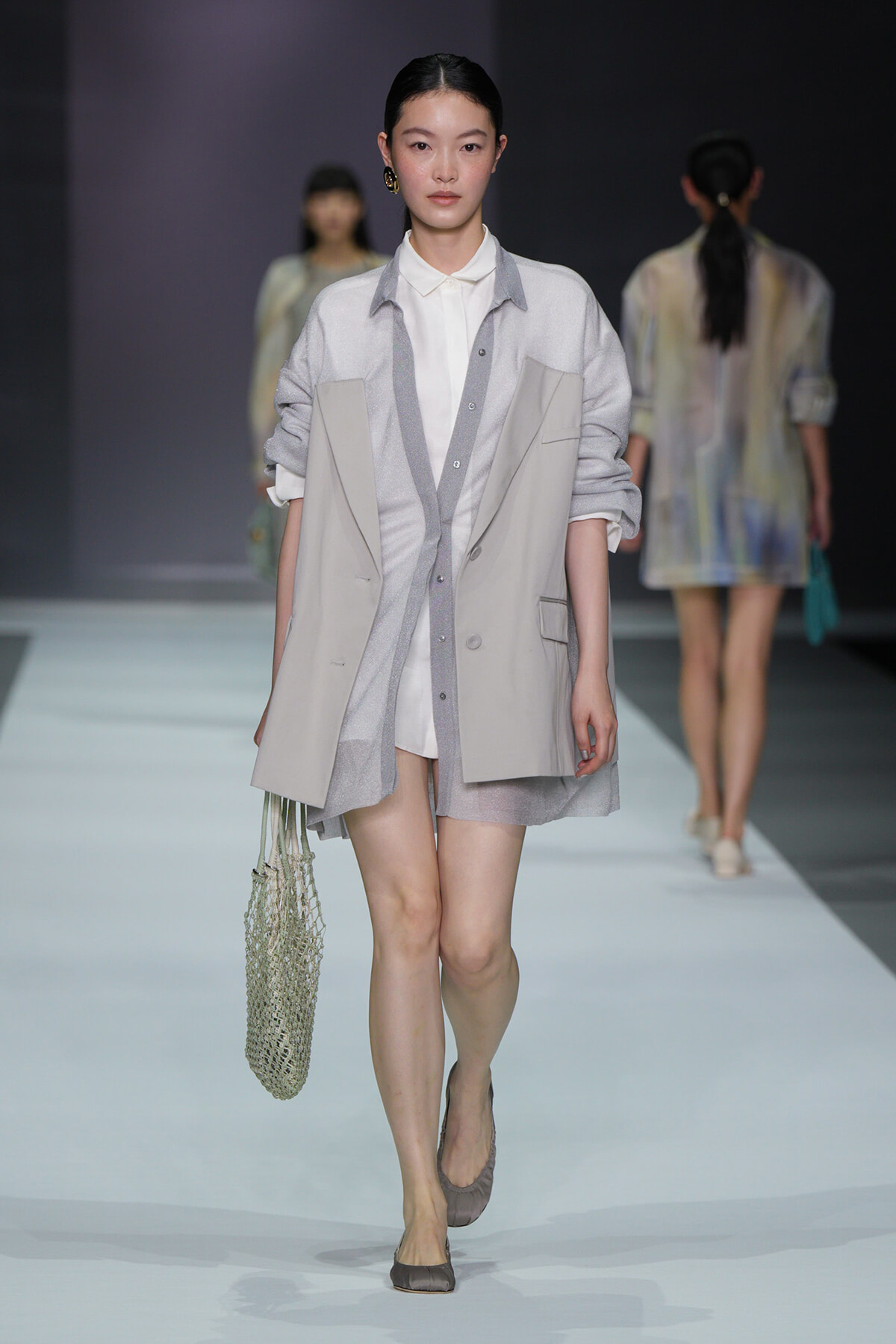 Model walking a fashion runway in a light gray layered outfit: white shirt, gray cardigan, oversized vest, beige flats, and a crochet net tote bag.
