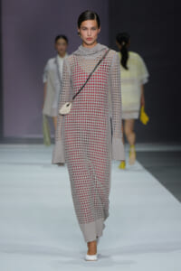 Model walking a runway in a gray perforated knit jumpsuit with a red underlayer, oversized cuffs, and a small white crossbody bag with white shoes.