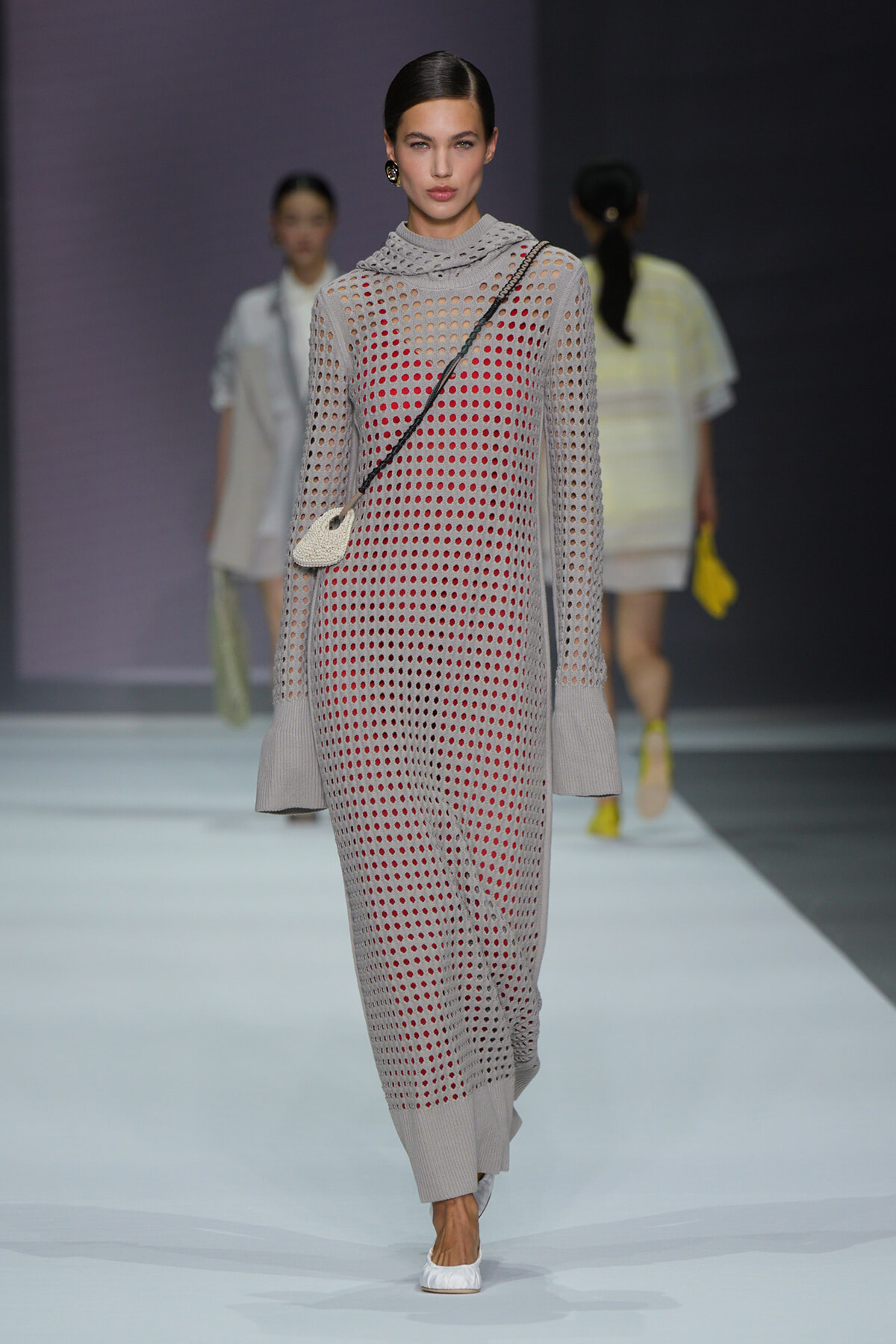Model walking a runway in a gray perforated knit jumpsuit with a red underlayer, oversized cuffs, and a small white crossbody bag with white shoes.