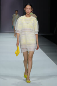 Model on a runway wearing a pale yellow striped, semi‑sheer tunic over matching shorts, holding a bright yellow textured clutch and wearing yellow flats.