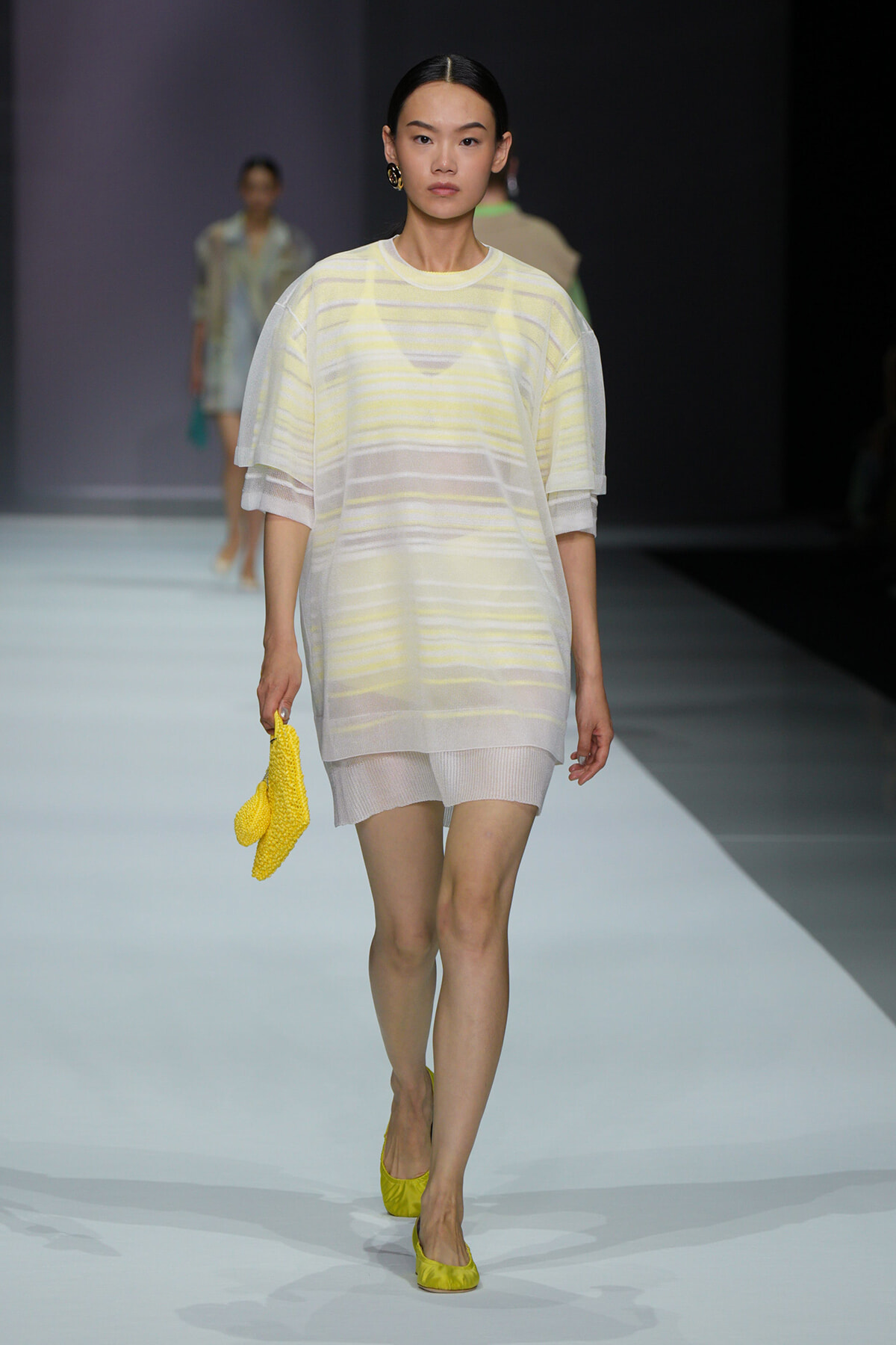 Model on a runway wearing a pale yellow striped, semi‑sheer tunic over matching shorts, holding a bright yellow textured clutch and wearing yellow flats.
