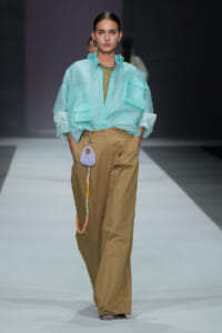 Model walking a runway in a loose turquoise button-up shirt with large pockets over an olive-green top and wide tan trousers; a small purple crocheted bag with a yellow-and-gray strap hangs from the belt loop.