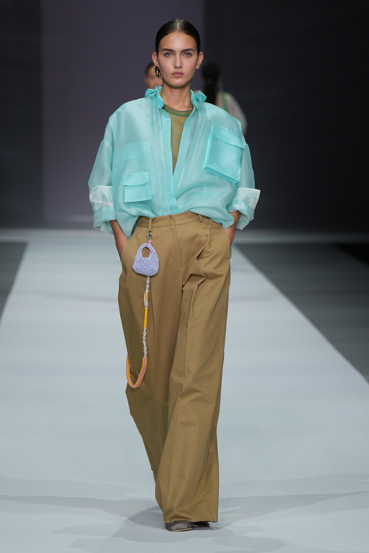 Model walking a runway in a loose turquoise button-up shirt with large pockets over an olive-green top and wide tan trousers; a small purple crocheted bag with a yellow-and-gray strap hangs from the belt loop.