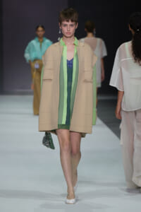 Model walks the runway wearing a beige oversized vest with exposed pockets over a lime-green and blue layered outfit, paired with white flats and a small beaded bag.