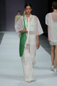 Model walking the fashion runway in a sheer white outfit with wide sleeves, carrying a bright green net tote bag on her shoulder.