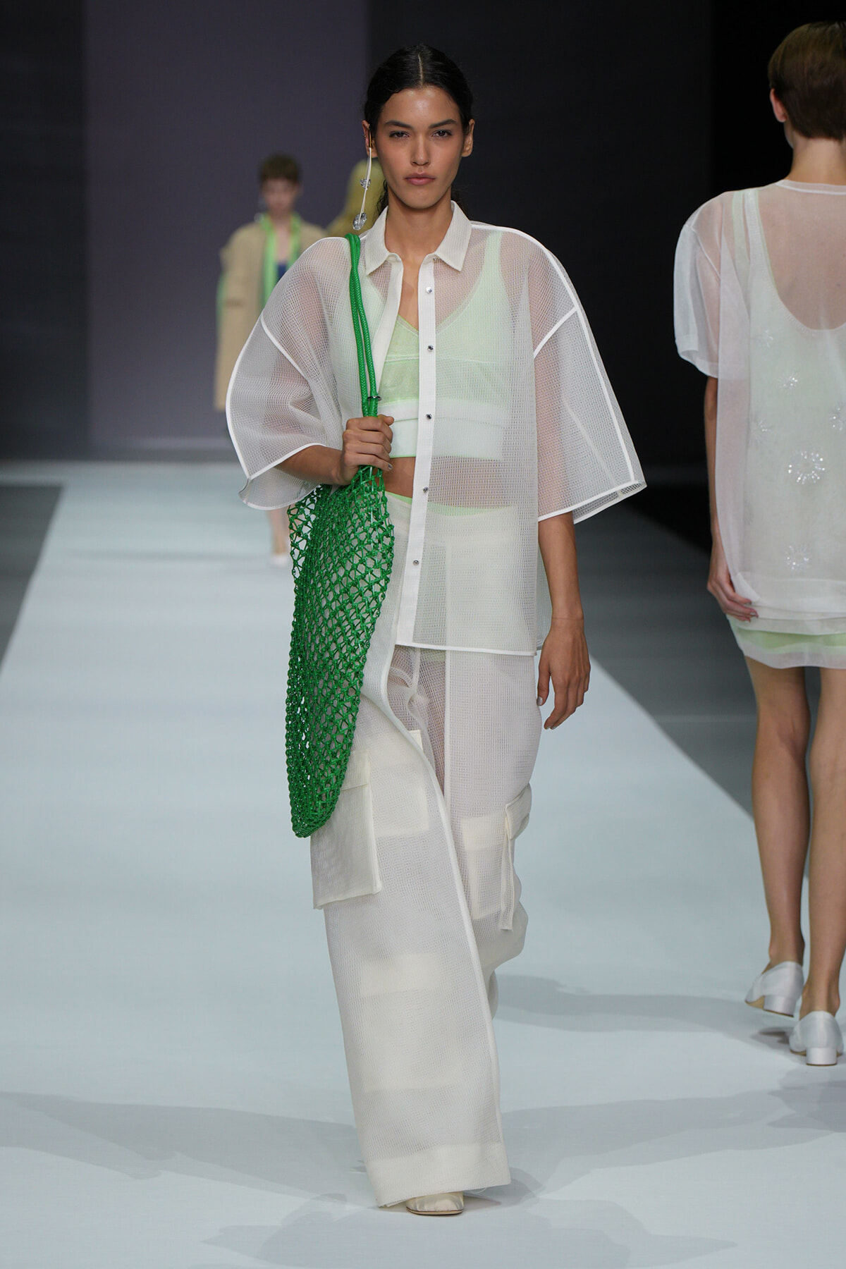Model walking the fashion runway in a sheer white outfit with wide sleeves, carrying a bright green net tote bag on her shoulder.