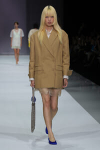 Model on a runway wearing an oversized tan blazer over a white shirt, sheer beige skirt, and blue heels, carrying a beaded clutch.