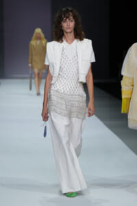 Runway model in a white layered ensemble: short vest over a beaded mesh top and wide-leg pants, with bright green shoes and a small blue accessory in hand.