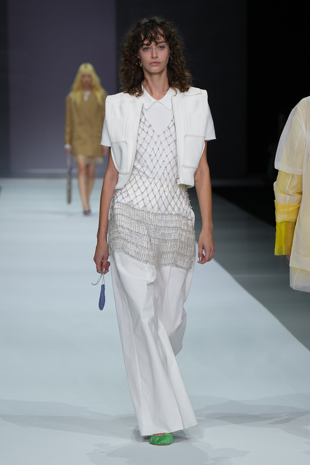 Runway model in a white layered ensemble: short vest over a beaded mesh top and wide-leg pants, with bright green shoes and a small blue accessory in hand.