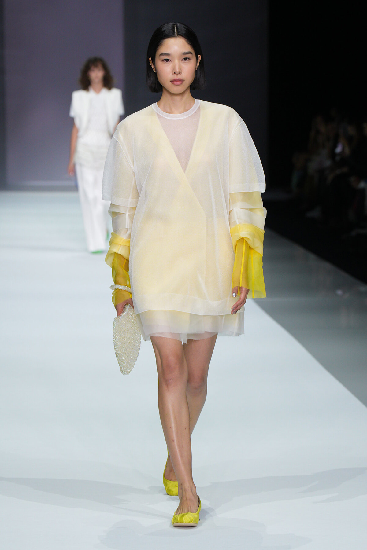 Model walks the runway in a pale yellow sheer layered tunic over white, with yellow accents, holding a beaded clutch and wearing yellow flats.