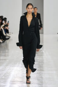 Model walks the runway in a black crinkled long-sleeve midi dress with a deep V neckline and high collar cuffs on the sleeves.