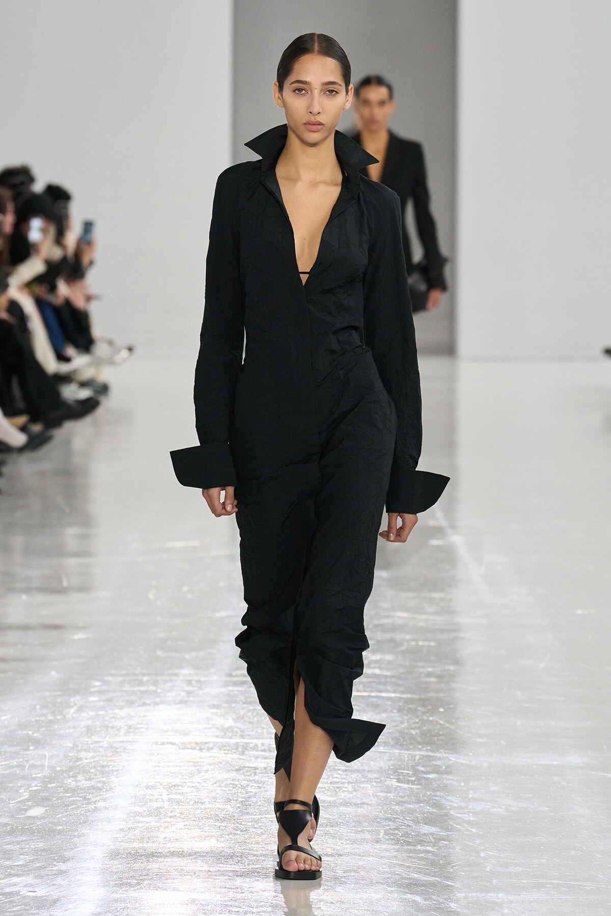 Model walks the runway in a black crinkled long-sleeve midi dress with a deep V neckline and high collar cuffs on the sleeves.