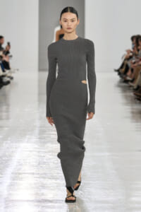 Model walks a grey ribbed knit maxi dress with side waist cutouts on a fashion show runway, spectators on both sides.