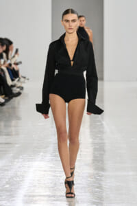 Model walking a runway in a black deep-V blouse with wide cuffs and high-waisted black shorts, black strappy heels.