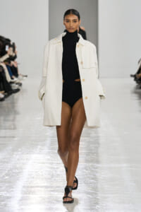 Model walking a fashion runway in an oversized white coat over a black ribbed turtleneck bodysuit with black sandals.