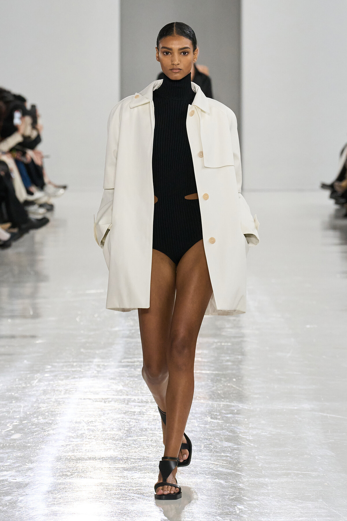 Model walking a fashion runway in an oversized white coat over a black ribbed turtleneck bodysuit with black sandals.