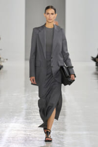 Female model walks a grey suit with blazer and matching skirt on a fashion runway, holding a black clutch and wearing black sandals.