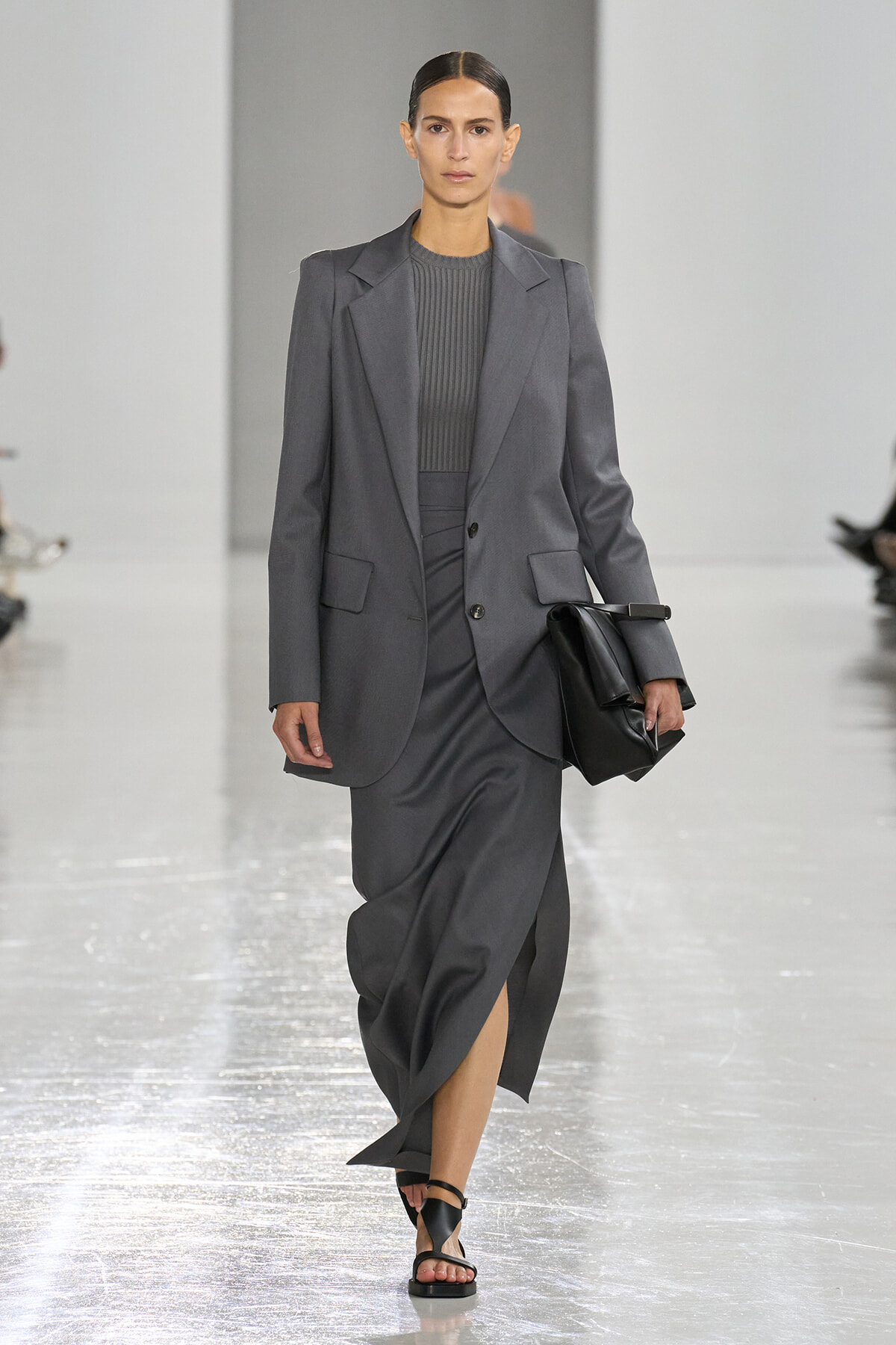 Female model walks a grey suit with blazer and matching skirt on a fashion runway, holding a black clutch and wearing black sandals.