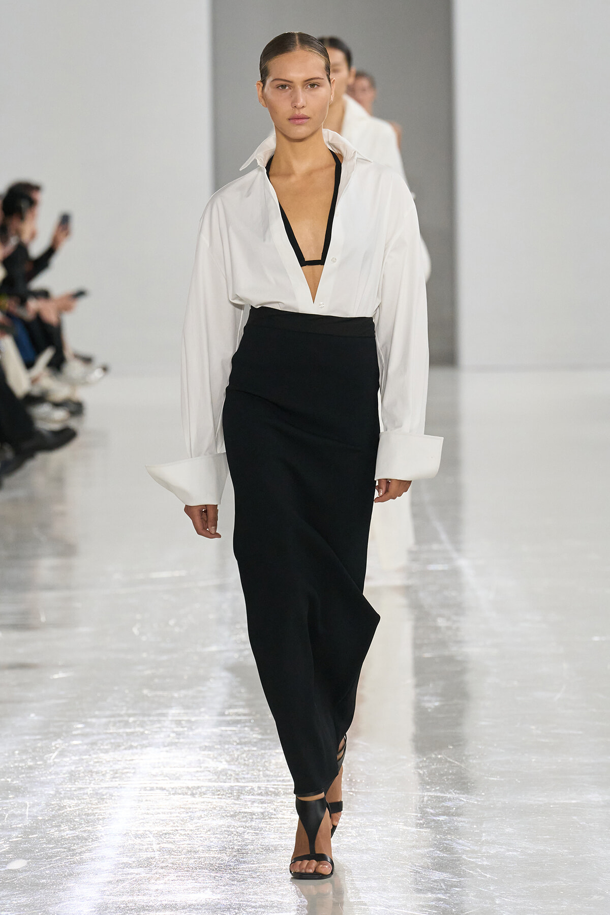 Model walks a fashion runway in a white oversized blouse with a plunging neckline and a black high-waisted skirt, paired with black sandals.