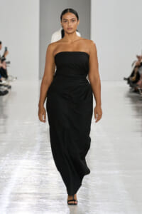 Model walking a runway in a strapless black gown, minimal jewelry, black sandals.