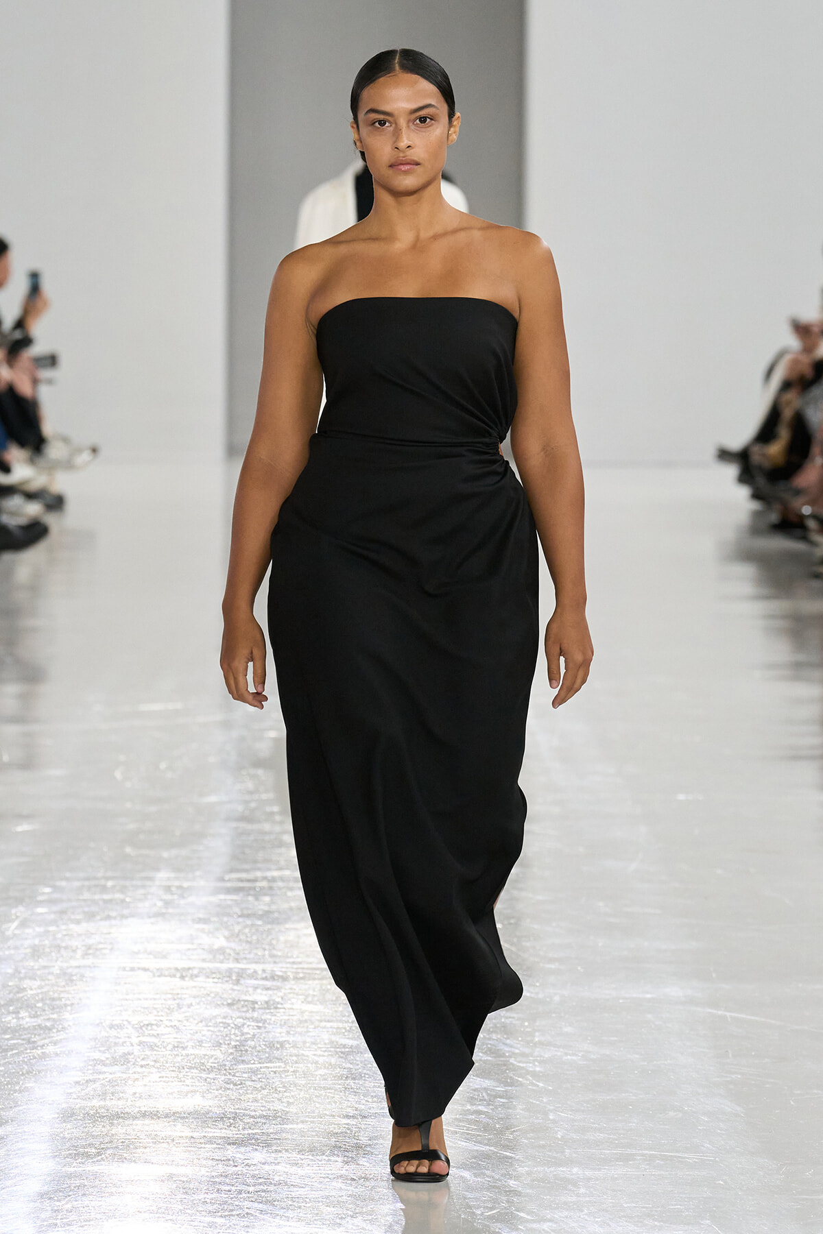Model walking a runway in a strapless black gown, minimal jewelry, black sandals.