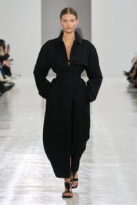 Model wearing a black belted trench-style dress with oversized sleeves walks the runway, wearing black sandals.
