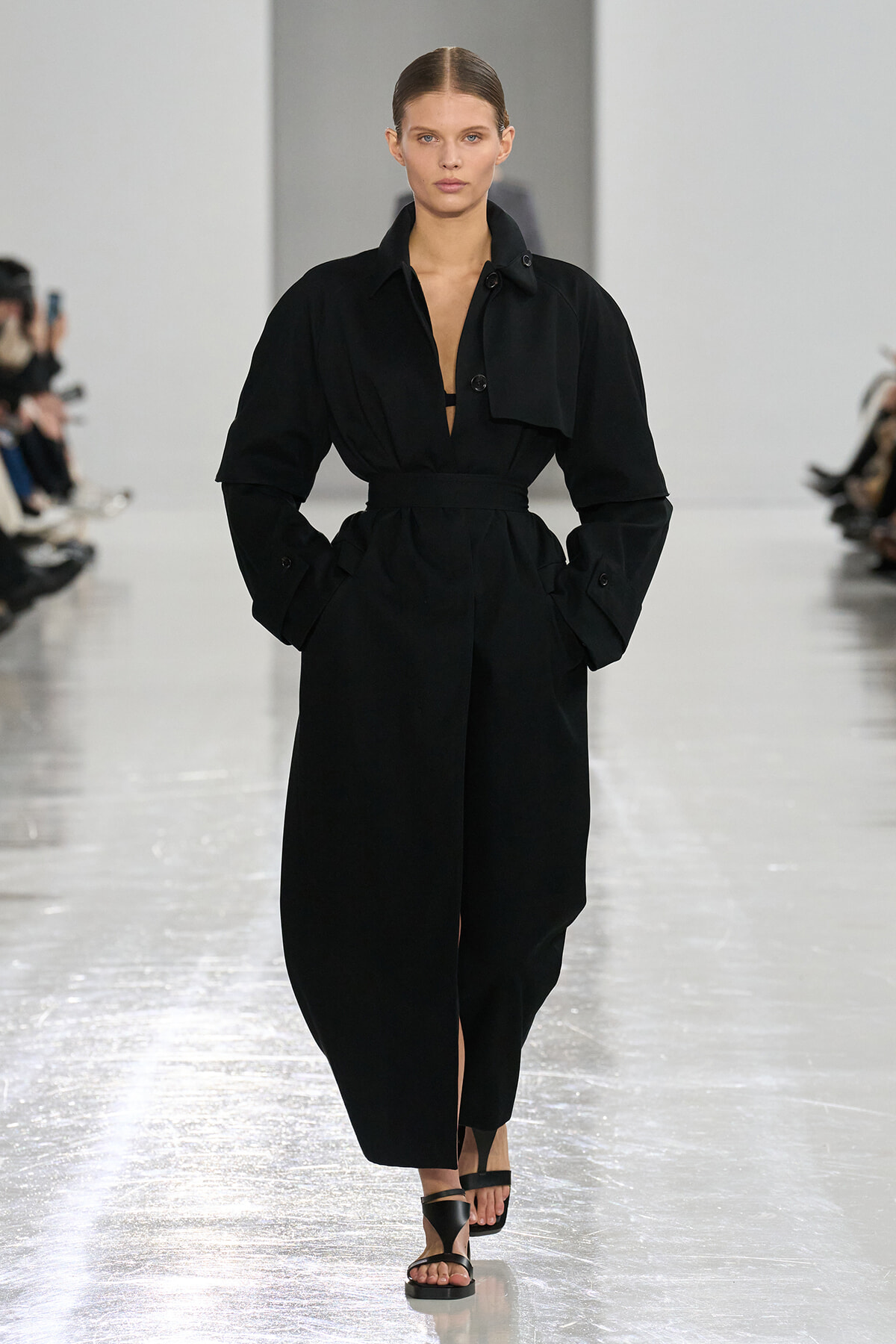 Model wearing a black belted trench-style dress with oversized sleeves walks the runway, wearing black sandals.
