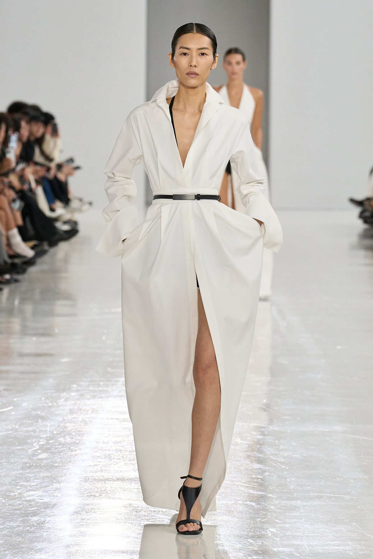 Model walking a fashion runway in a long white trench coat belted at the waist, black sandals, audience on the left watching.