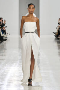 Model walks the runway in a strapless white gown with a high slit, cinched with a black belt and black heeled sandals.