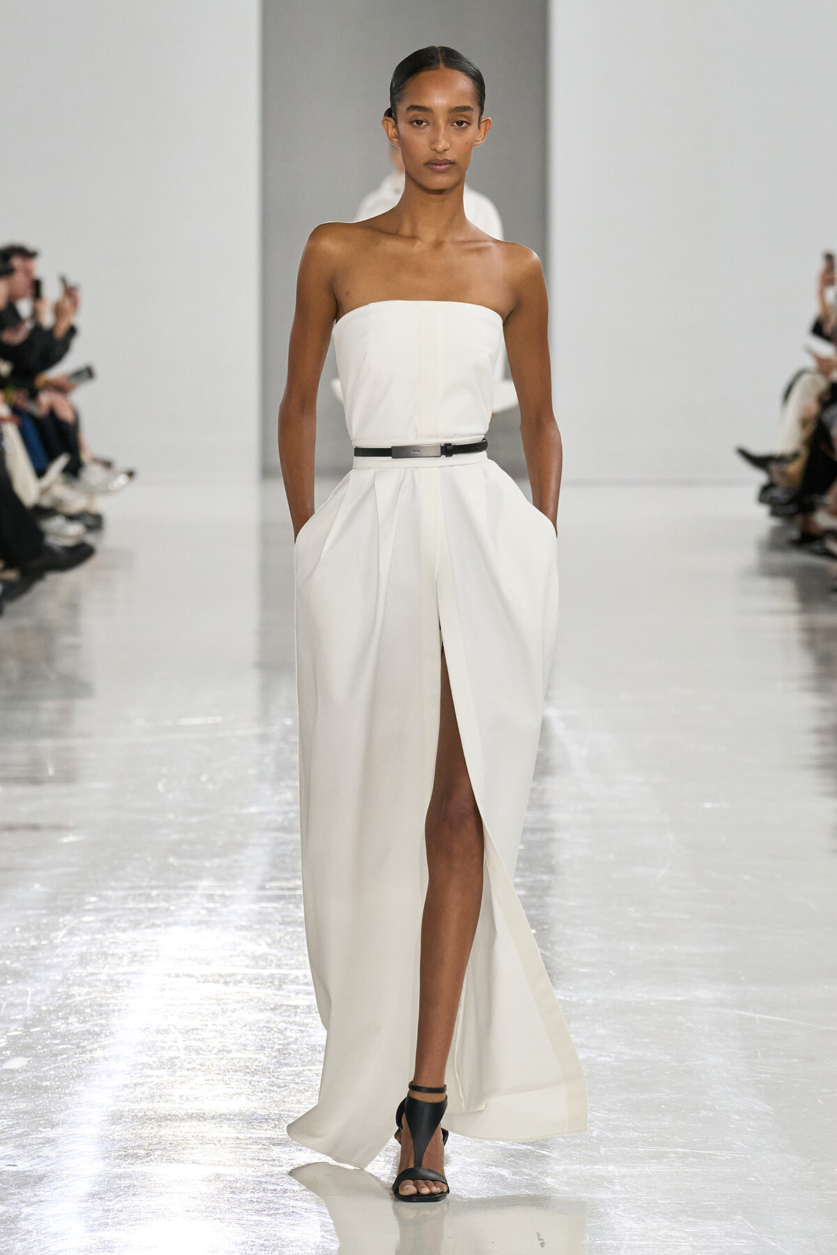 Model walks the runway in a strapless white gown with a high slit, cinched with a black belt and black heeled sandals.