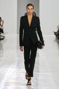 Model on a fashion runway wearing a black tailored blazer and matching trousers, carrying a small black clutch.