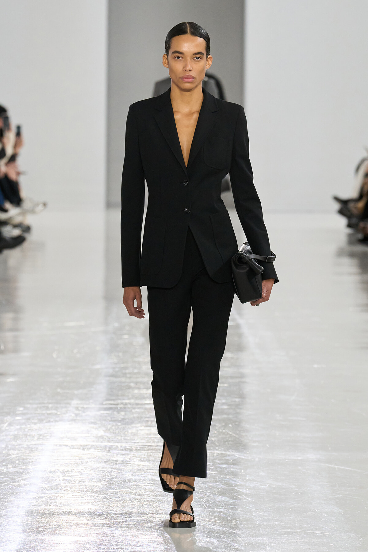 Model on a fashion runway wearing a black tailored blazer and matching trousers, carrying a small black clutch.