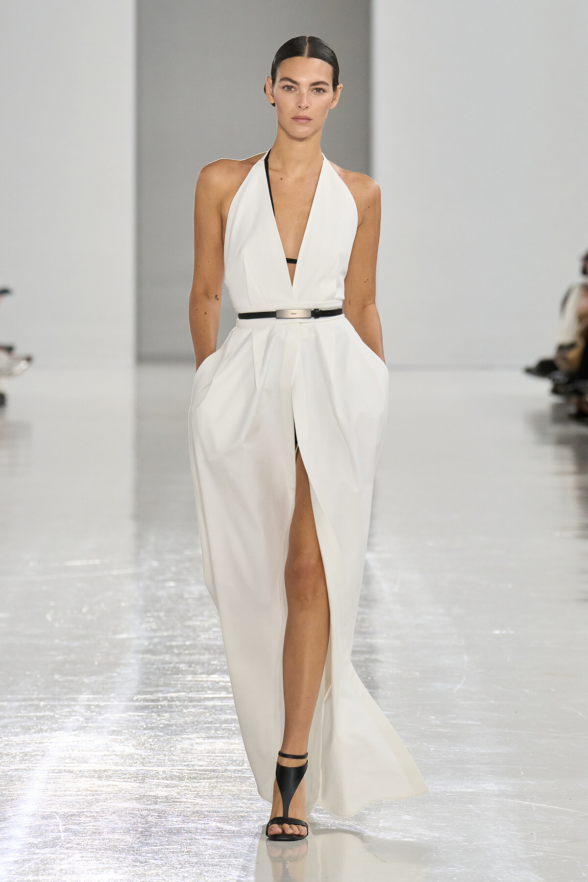Runway model in a white halter-neck gown with a deep V, black belt, and high front slit.