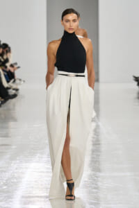 Model on a white runway wearing a black halter top and white high-waist skirt with a front slit and black belt.