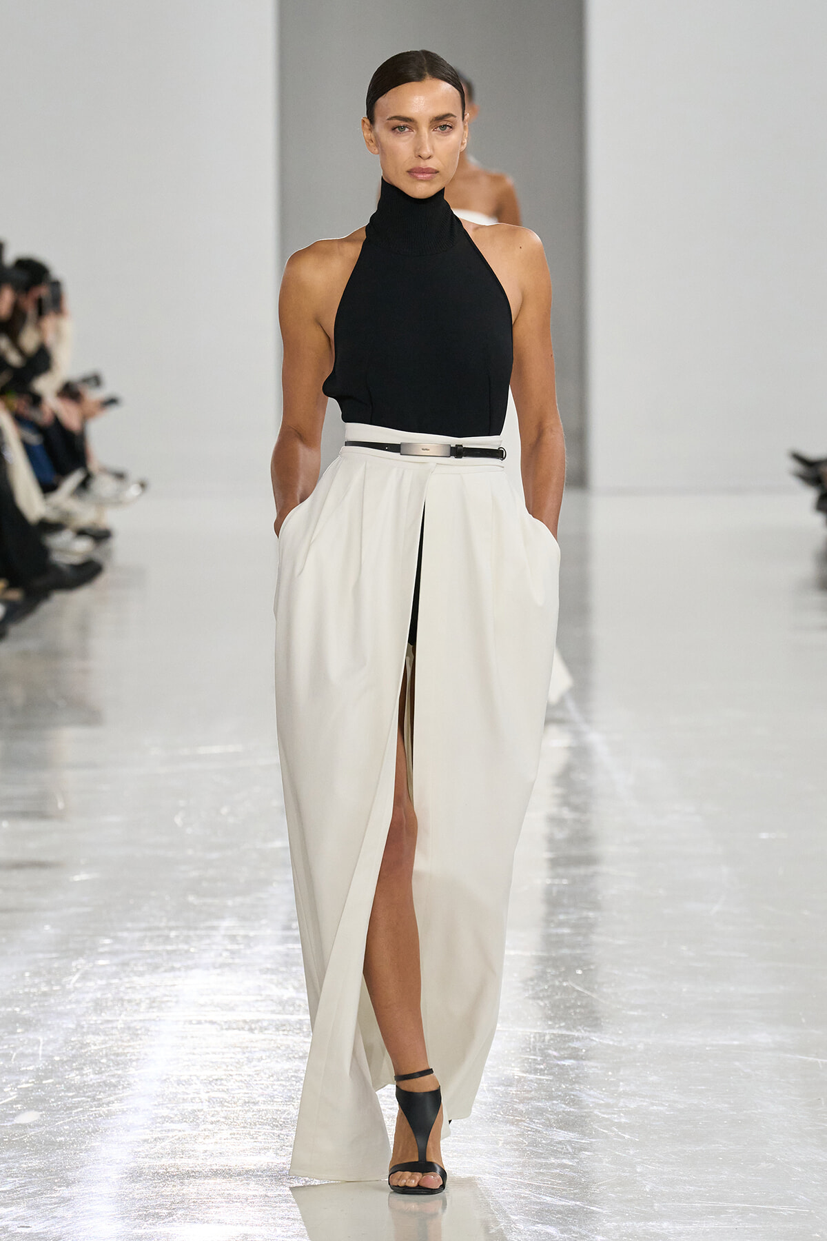 Model on a white runway wearing a black halter top and white high-waist skirt with a front slit and black belt.